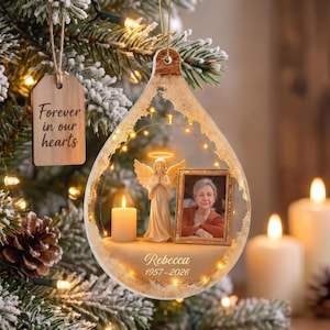 May include: A teardrop-shaped memorial ornament featuring a central angel figurine, a lit candle, and a framed photograph of a person. The ornament includes the name Rebecca and the dates 1957-2026, with a wooden tag reading Forever in our hearts.