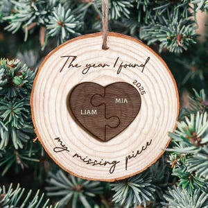 May include: A wooden Christmas tree ornament with a natural wood grain. A dark brown heart-shaped puzzle is in the centre, with the names "LIAM" and "MIA" on each piece. The text "The year I found 2025 my missing piece" is written around the ornament.