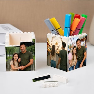 May include: Two white block-style pen holders with family photos. One holds colorful markers. The other is empty. Loose blocks are in front. The pen holders are on a white surface.