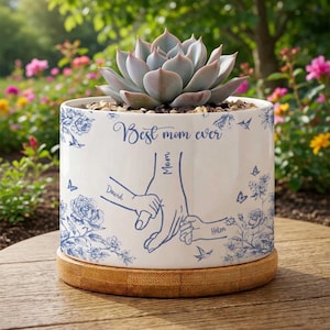 May include: A white ceramic planter with a blue floral design and the text "Best mom ever." The planter holds a succulent plant and sits on a wooden base. The design includes illustrations of hands holding each other, with the names "David" and "Helen."
