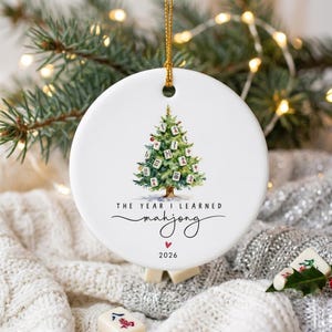 May include: A round white ceramic ornament featuring a watercolor illustration of a Christmas tree decorated with mahjong tiles. The text reads THE YEAR I LEARNED mahjong above a small red heart and the year 2026. It hangs from a gold cord.