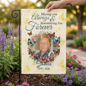 Può includere: Una bandiera da giardino rettangolare con il ritratto di una persona anziana con gli occhiali, circondato da una ghirlanda di farfalle colorate. Il testo recita: Missing you Always & Remembering you Forever, Shane, 1959-2026. Fondo color crema.