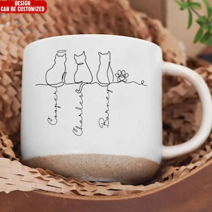 May include: White ceramic mug with a brown base, featuring a line drawing of three cats and a paw print. The names "Cooper", "Charles", and "Barney" are written below the cats. The top left corner reads "DESIGN CAN BE CUSTOMIZED".