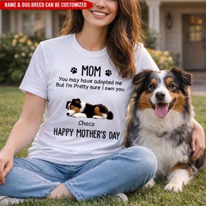 May include: White t-shirt with the text "MOM" and "HAPPY MOTHER'S DAY". The shirt features a graphic of a sleeping dog and the phrase "You may have adopted me But I'm Pretty sure I own you". A dog is sitting next to the person wearing the shirt.