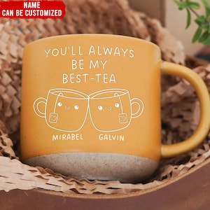 May include: Mustard yellow ceramic mug with a light brown base and a curved handle. The mug features the text "YOU'LL ALWAYS BE MY BEST-TEA" and two smiling teacup illustrations. The names "MIRABEL" and "GALVIN" are printed below the teacups.