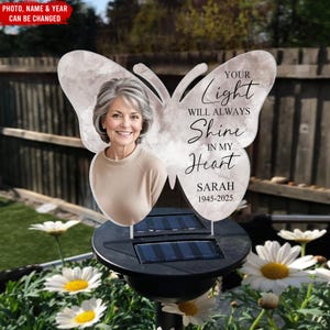 Pode incluir: Uma estaca de jardim movida a energia solar com um design de borboleta e uma foto de uma mulher. O texto diz "Your Light Will Always Shine In My Heart" com o nome "SARAH" e as datas "1945-2025". O topo tem formato de borboleta.