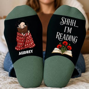 May include: A pair of black and sage green socks. The left sock features an illustration of a person reading a book while wrapped in a red patterned blanket, with the name AUDREY printed below. The right sock displays the text SHHH... I'M READING above an open book with red poppy flowers.