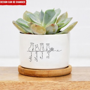 May include: A white ceramic plant pot with a succulent. The pot has a black line drawing of four cats with halos and names below. It sits on a round wooden coaster. The text "DESIGN CAN BE CHANGED" is at the top.