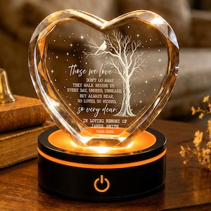 May include: A heart-shaped clear crystal plaque featuring an etched tree, a bird, and the text: Those we love don't go away they walk beside us every day, unseen, unheard, but always near, so loved, so missed, so very dear. In loving memory of James Smith 1948-2026. The crystal sits on a round black light base with an orange power button.