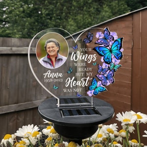 May include: A heart-shaped memorial plaque with a photo of a woman and the text "Your Wings Were Ready But My Heart Was Not". The plaque is decorated with blue and purple butterflies and is mounted on a black solar-powered stake.