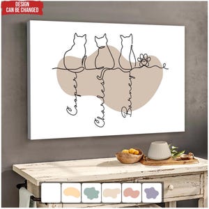 May include: A canvas print with a minimalist line drawing of three cats and a paw print. The names "Cooper", "Charles", and "Barney" are written below the cats. The background includes a neutral-toned shape. The text "DESIGN CAN BE CHANGED" is in the upper left corner.