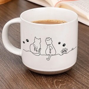 May include: White ceramic coffee mug with a brown base, filled with coffee. The mug features a black line drawing of two cats, paw prints, and the names "Jett" and "Coco". A flower design is also present.