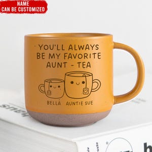 Op de afbeelding: Een gele keramische mok met een bruine basis. De mok heeft een zwarte afbeelding van twee theekopjes met gezichten en de tekst "You'll always be my favorite aunt - tea". De tekst "Bella Auntie Sue" staat onder de theekopjes.