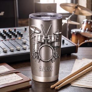 May include: A metallic silver tumbler featuring a raised, embossed design of a complete drum kit with cymbals and drumsticks. The name ALEX is engraved in capital letters near the base. The tumbler has a clear plastic lid and a smooth finish.