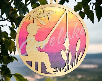 May include: A circular wooden ornament with a silhouette of a person fishing. The background features a watercolour sunset in shades of yellow, pink, and purple. The text "Love Lake" is visible. The ornament has a hole for hanging.