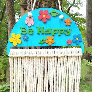 May include: A turquoise blue wooden half-circle sign with the text "Be Happy" in green letters. The sign is decorated with colorful flowers and leaves. The sign is hanging from a white macrame wall hanging.