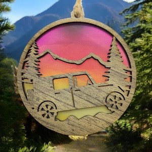 Puede incluir: Un adorno de madera con forma de jeep conduciendo por un paisaje montañoso con una puesta de sol en el fondo. El adorno está pintado con un degradado de rosa, naranja y amarillo.