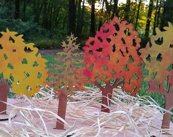 May include: Four wooden cutout trees in various autumn colours. The trees are standing on a bed of straw-like material. The trees are painted in shades of yellow, orange, red, and brown.