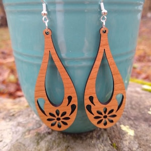 May include: A pair of wooden earrings with a teardrop shape and a floral cutout design. The earrings are a light brown color and have silver hooks.