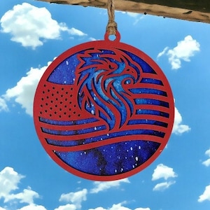 Puede incluir: Adorno de madera rojo con un diseño de bandera estadounidense azul y blanca y una silueta de águila. El adorno está colgado de una cuerda contra un cielo azul con nubes blancas.