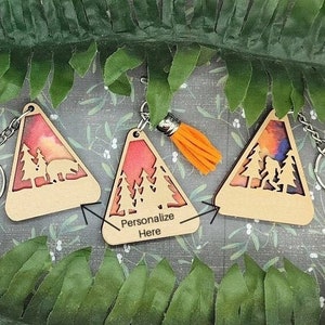 May include: Wooden triangle keychains with a forest and sunset design. One keychain has the text "Personalize Here". Each keychain has a metal chain and an orange tassel.