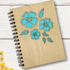 May include: A wooden spiral-bound notebook with a floral design. The cover features three turquoise flowers and leaves, laser-cut into the light-colored wood. The notebook's spiral binding is black.