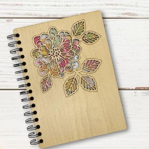 May include: A wooden notebook with a spiral binding, featuring a laser-cut floral design on the cover. The flower is filled with colourful watercolour-style patterns. The notebook is made of light-coloured wood and has a black spiral binding.