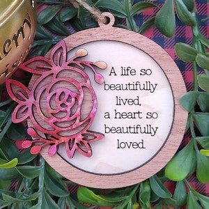 Puede incluir: Un adorno navideño de madera con un diseño floral rojo y rosa y el texto "Una vida tan bellamente vivida, un corazón tan bellamente amado."