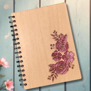 May include: A wooden spiral-bound notebook with a floral design. The cover is light brown with a laser-etched floral arrangement in shades of purple. The spiral binding is black.