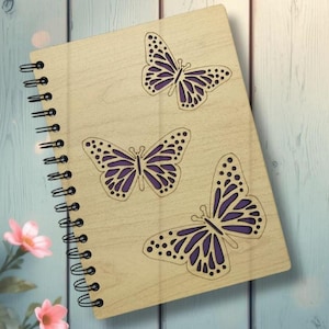 May include: A wooden notebook with a spiral binding, featuring three laser-cut butterfly designs with purple accents. The notebook has a light wood grain finish and is set against a blue and white background.