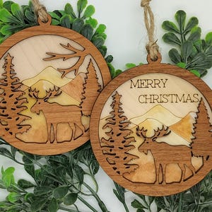 May include: Two round wooden Christmas ornaments with a nature scene featuring a moose, trees, and mountains. One ornament has the text "MERRY CHRISTMAS". Each ornament has a jute string for hanging.