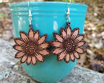 Könnte beinhalten: Ein Paar Ohrringe aus Holz in Form von Sonnenblumen. Die Ohrringe sind hellbraun und haben ein detailliertes Design. Die Ohrringe hängen an silbernen Haken.