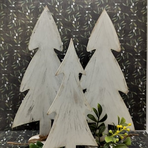 May include: Three white wooden Christmas trees of varying sizes, with a distressed paint finish, stand on a dark background. The trees are arranged in a staggered formation, with the largest tree in the back and the smallest tree in the front.