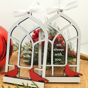 May include: Two white wooden ornaments shaped like arched windows, each with a red cardinal and the text "We are always with you" and "I am always with you." The ornaments have white ribbons for hanging.