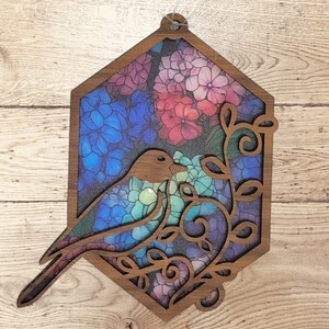 Pu&ograve; includere: Un ritaglio di legno di un uccello con ali in vetro colorato blu, verde e viola, appollaiato su un ramo con fiori colorati. Il ritaglio ha la forma di un esagono e ha un foro nella parte superiore per appenderlo.