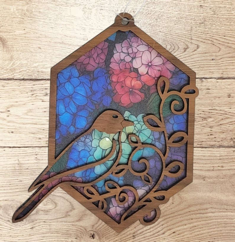 Pu&ograve; includere: Un ritaglio di legno di un uccello con ali in vetro colorato blu, verde e viola, appollaiato su un ramo con fiori colorati. Il ritaglio ha la forma di un esagono e ha un foro nella parte superiore per appenderlo.