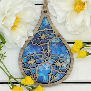 May include: A wooden teardrop-shaped ornament with a blue stained glass design featuring three stylized flowers. The ornament is surrounded by white and yellow flowers.