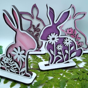 May include: Three wooden bunny decorations with a layered design. The bunnies are painted in shades of pink and purple with white floral accents. The bunnies are standing on white bases.