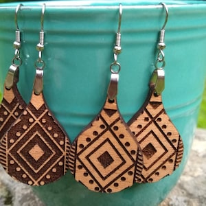 Puede incluir: Tres pares de pendientes de madera con un patrón geométrico. Los pendientes son marrones con un patrón geométrico marrón claro y tienen ganchos plateados.