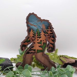 May include: A wooden cutout of Bigfoot walking through a forest with an owl perched on a branch. The cutout is painted in shades of brown, green, and blue.