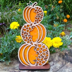 Może przedstawiać: Drewniany wycinany wzór trzech ułożonych na sobie dyni z misternymi wzorami kwiatowymi. Dynie są pomarańczowe, a kwiaty białe. Wzór znajduje się na brązowej podstawie i stoi na kamieniu.
