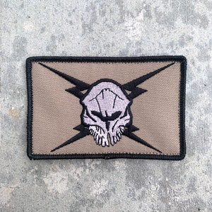 May include: A tan embroidered patch with a black border. The patch features a skull with lightning bolts behind it.