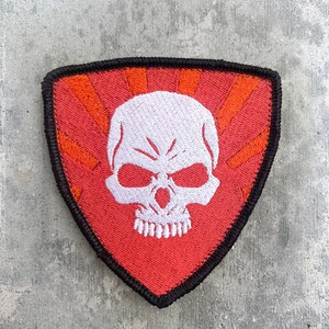 Könnte beinhalten: Ein schildförmiger, bestickter Aufnäher mit einem weißen Totenkopf auf rotem Hintergrund, der von orangefarbenen Sonnenstrahlen umgeben ist. Der Aufnäher ist mit einem dicken, schwarz gestickten Rand um den gesamten Umfang versehen.