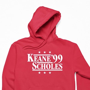 May include: A red hooded sweatshirt with the text "Keane '99 Scholes" in white, with three stars above and below the text. The sweatshirt has a front pocket and a drawstring hood.