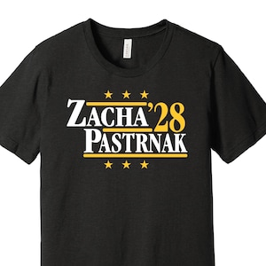 以下が含まれることがあります： 白と黄色の「Zacha '28 Pastrnak」の文字が入った黒いTシャツ。デザインには、テキストの上と下に星が描かれています。クルーネックのTシャツです。
