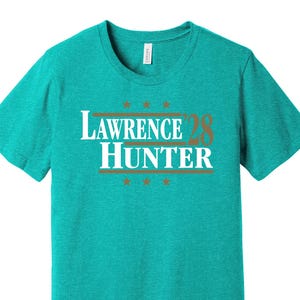 May include: A teal short-sleeve t-shirt with the text "LAWRENCE HUNTER 28" in white and brown. The text is centered on the chest, with stars above and below the text. The shirt is made of a soft material.