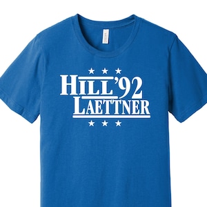 Peut inclure: Un t-shirt bleu roi avec le texte blanc "Hill '92 Laettner", avec trois étoiles au-dessus et en dessous. Le t-shirt est fait d'une matière douce et a un col rond.