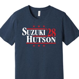 Könnte beinhalten: Marineblaues T-Shirt mit dem Text "SUZUKI '28 HUTSON" in Weiß und Rot. Das Design umfasst rote Sterne über und unter dem Text. Das Shirt hat einen Rundhalsausschnitt und kurze Ärmel.
