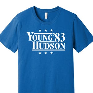 May include: A royal blue t-shirt featuring the white text "Young '83 Hudson", with three stars above and below. The t-shirt has a crew neck design.