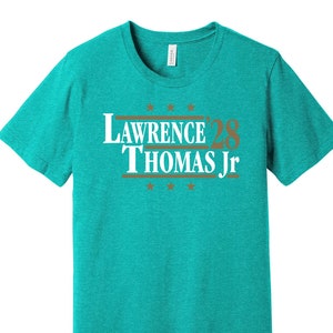 May include: A turquoise blue t-shirt with a white print that says "Lawrence '28 Thomas Jr." in a vintage political campaign style.